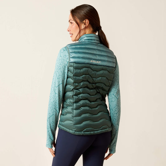 Rear view of a young woman wearing an ariat green two-tone puffer gilet and base layer with stand up collar, standing against a neutral background