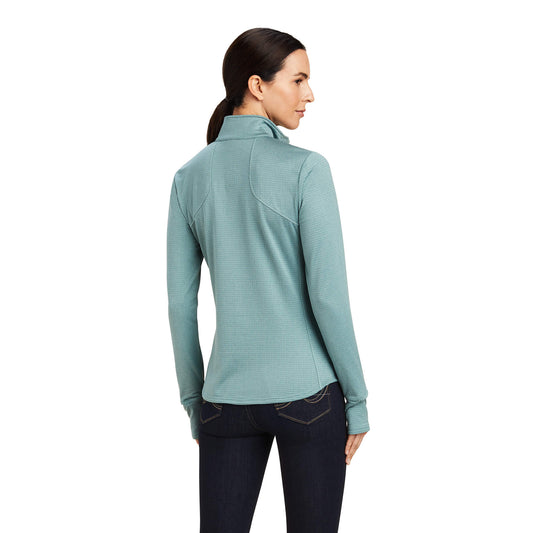 Back view of a young woman wearing a light blue ariat long-sleeve zip-up base layer with thumb holes, standing against a white background
