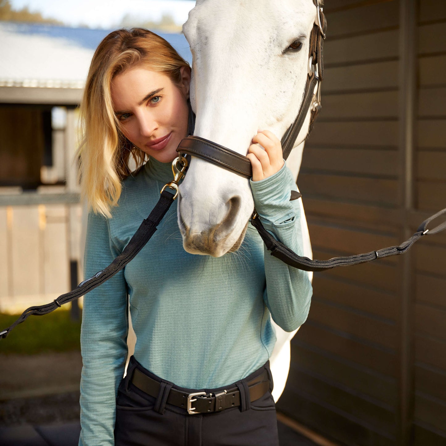 Young woman wearing a light blue ariat long-sleeve zip-up base layer with thumb holes and a smart-watch window, standing next to and holding a white horse