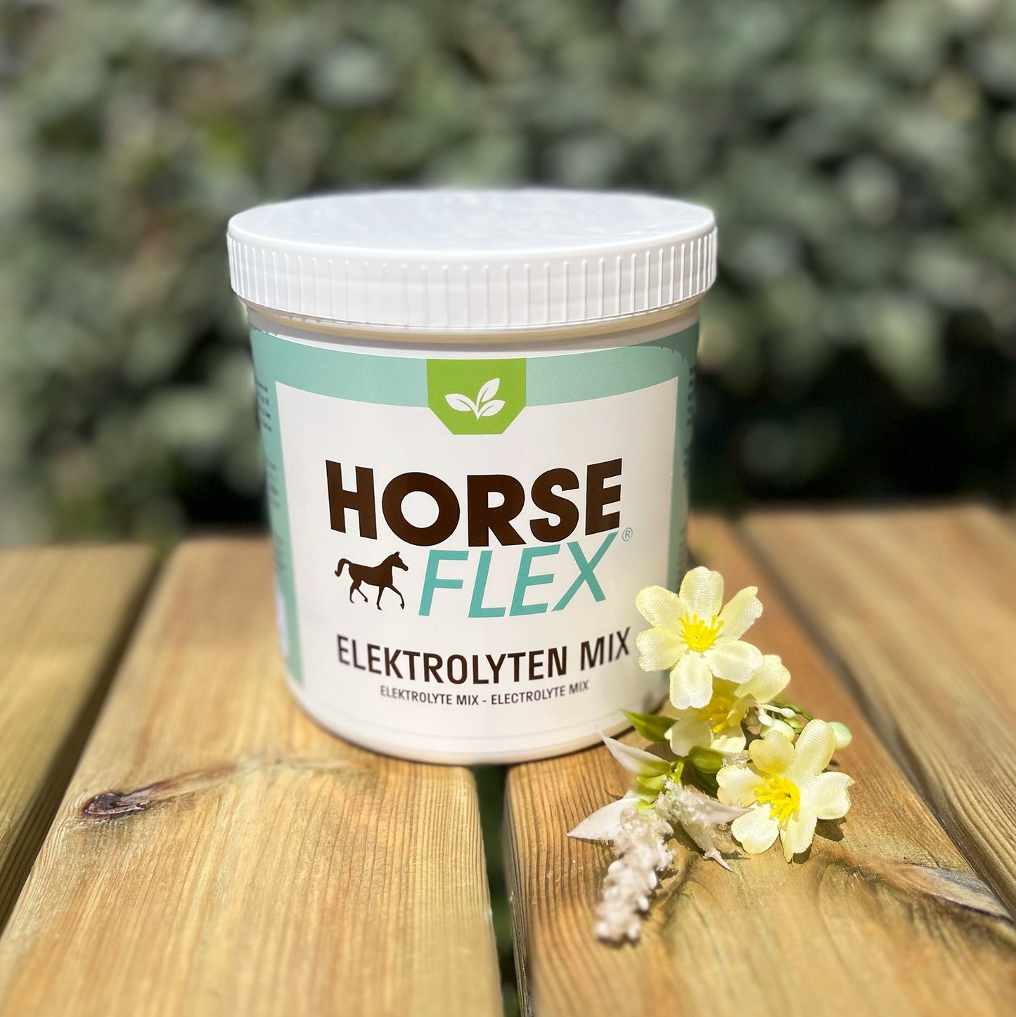 A pot of Horseflex electrolyte mix powder on top of a wooden table against a green bush background, next to some flowers